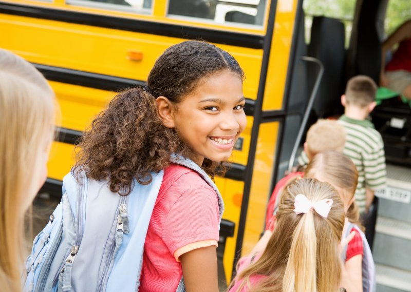Girl turning around to face camera with a smile in a line of children getting on a school bus